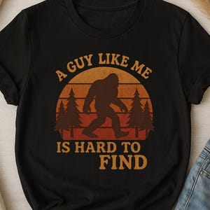 Peut inclure: T-shirt noir avec un motif de Bigfoot dans un coucher de soleil. Le texte dit "A guy like me is hard to find". Le t-shirt est associé à un jean bleu.