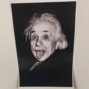 May include: Black and white portrait print of Albert Einstein sticking out his tongue. The image shows Einstein with his eyes wide and his tongue extended, wearing a dark suit. The print is on white paper.