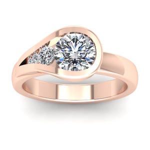 Może przedstawiać: Pierścionek z różowego złota z dużym, okrągłym, przezroczystym kamieniem szlachetnym w oprawie z ramką, z trzema mniejszymi kamieniami szlachetnymi osadzonymi w zakrzywionym kanale. Pierścionek ma gładką, wypolerowaną obrączkę i nowoczesny, elegancki design.