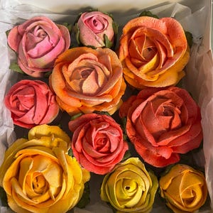 May include: A white box containing nine buttercream roses in shades of pink, orange, and yellow. The roses have green leaves and are arranged in a visually appealing pattern. The box is lined with white paper.