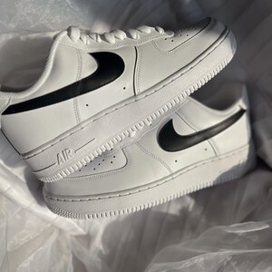 May include: Pair of white sneakers with black swoosh logos. The shoes have white laces and soles, with the word "AIR" printed on the side. The sneakers are displayed on a white, textured surface.