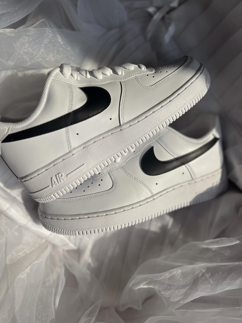 May include: Pair of white sneakers with black swoosh logos. The shoes have white laces and soles, with the word "AIR" printed on the side. The sneakers are displayed on a white, textured surface.