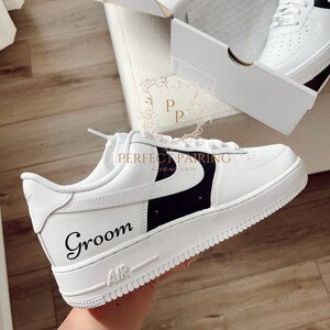 May include: White leather trainers with a black swoosh and the word "Groom" written in black script. The trainers have a white sole with black trim.