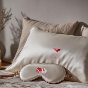 May include: A cream-colored silk eye mask and pillow with embroidered designs. The eye mask features two red dice, and the pillow has a pink cocktail glass. The items are on a bed with neutral-toned pillows.