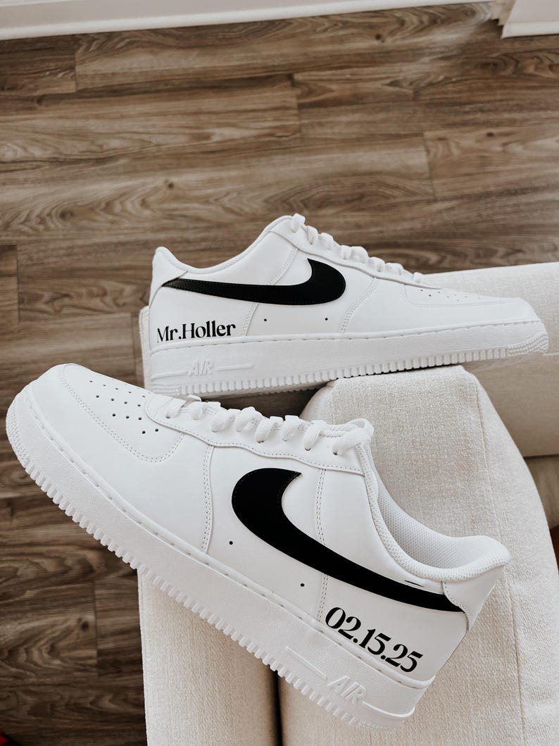 May include: A pair of white sneakers with black accents, including the Nike swoosh logo. The text "Mr. Holler" is printed on one shoe, and "02.15.25" on the other. The shoes are displayed on a white surface.