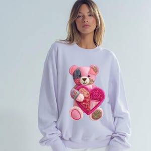 Op de afbeelding: Een lichtpaarse sweatshirt met een patchwork teddybeer die een roze hart vasthoudt. De beer heeft roze, zwarte en gouden accenten. De sweatshirt heeft een relaxte pasvorm, lange mouwen en een ronde hals.