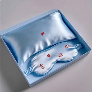 May include: A light blue silk sleep set in a matching box. The set includes a small pillow and an eye mask. The pillow has a red bow embroidered on it, and the eye mask has red apple designs.