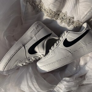 May include: A pair of white leather sneakers with black swoosh logos. The shoes are placed on a white, textured surface, with a decorative beaded embellishment in the background. The sneakers have a classic, sporty design.