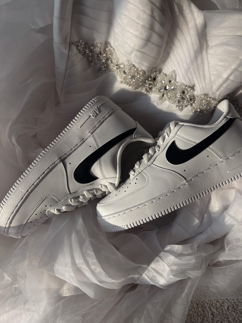 May include: A pair of white leather sneakers with black swoosh logos. The shoes are placed on a white, textured surface, with a decorative beaded embellishment in the background. The sneakers have a classic, sporty design.