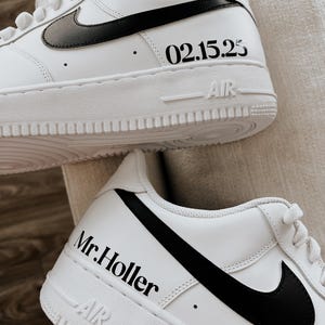 May include: White sneakers with black swooshes and soles. The text "02.15.25" is printed on the side of one shoe, and "Mr. Holler" is printed on the side of the other shoe. The word "AIR" is printed on the sole.
