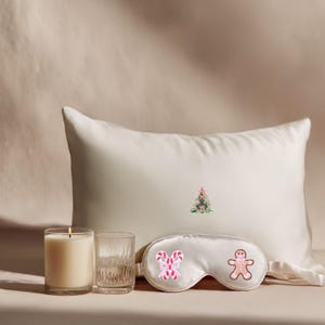 May include: A cream-colored pillow with a small embroidered Christmas tree. A white eye mask with candy cane and gingerbread man designs is in front of the pillow. A lit candle and a glass are on the left.