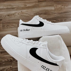 May include: A pair of white sneakers with black accents, including the Nike swoosh logo. The text "Mr. Holler" is printed on one shoe, and "02.15.25" on the other. The shoes are displayed on a white surface.