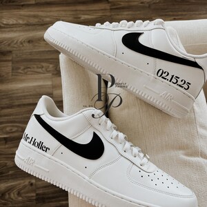 May include: A pair of white sneakers with black swoosh logos. One shoe has "Mr. Holler" printed on the side, and the other has "02.15.25". The shoes are on a cream-colored surface.