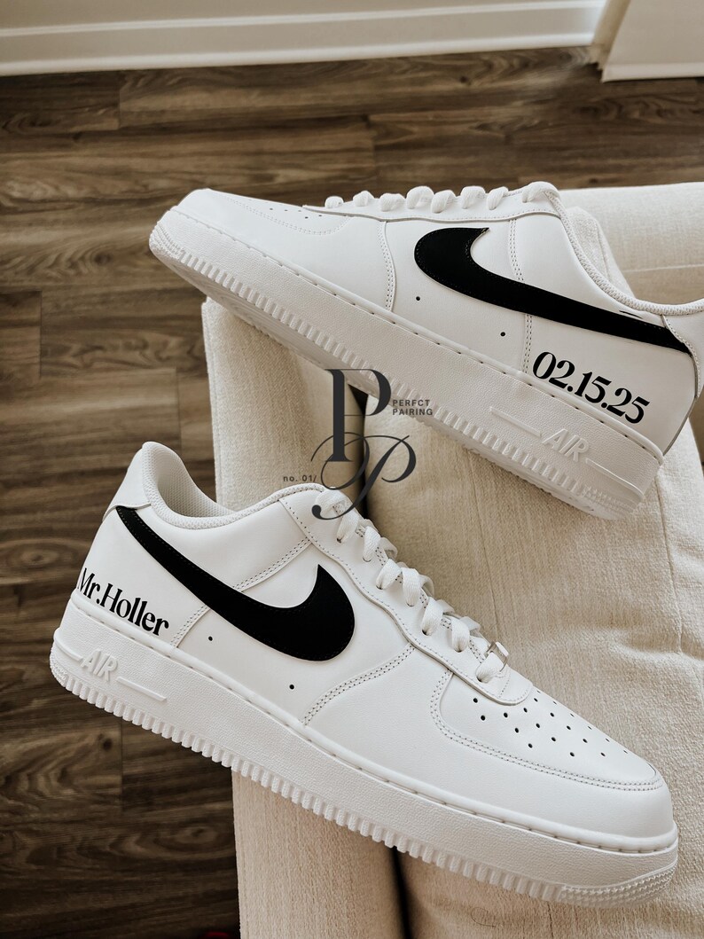 May include: A pair of white sneakers with black swoosh logos. One shoe has "Mr. Holler" printed on the side, and the other has "02.15.25". The shoes are on a cream-colored surface.