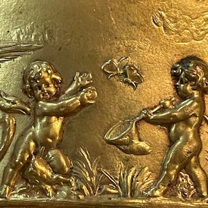 May include: A gold-toned metal box with a decorative design featuring two cherubs. One cherub is holding a net and the other is reaching for a butterfly. The box has a detailed floral border.