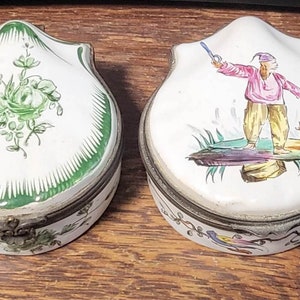 May include: Two antique porcelain trinket boxes with hinged lids. One box features a green floral design, while the other depicts a figure in a pink shirt and yellow pants holding a sword.