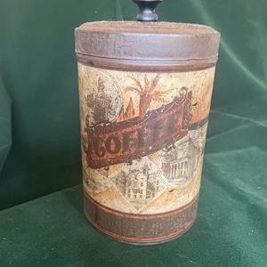 May include: Vintage cylindrical coffee tin with a lid and a small knob. The tin features a brown and beige design with the word "COFFEE" in large letters, along with illustrations of buildings and a vehicle. The tin is made of metal and has a rustic appearance.