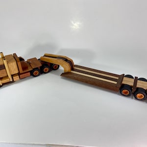 May include: A wooden toy truck and trailer set. The truck cab is made of various wood tones, with black wheels and orange hubs. The trailer is a dark wood with lighter wood stripes and black wheels with orange hubs.
