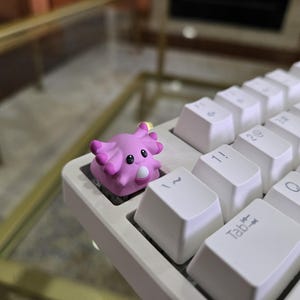 May include: A pink axolotl-shaped artisan keycap featuring a smooth matte finish, black eyes, and a white snout, positioned on the top left corner of a white mechanical keyboard with standard alphanumeric keys and a tab key visible.