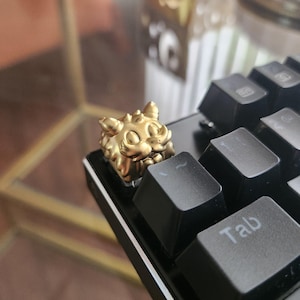 May include: A metallic gold-colored artisan keycap shaped like a stylized creature with pointed ears and a textured face, installed on a black mechanical keyboard next to a key labeled with the text Tab in white lettering.