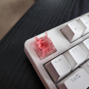 May include: A translucent pink resin artisan keycap shaped like a horned demon mask, installed on a white mechanical keyboard. The keycap features detailed facial features including horns, a wide mouth, and defined eyes, contrasting with the white keys.
