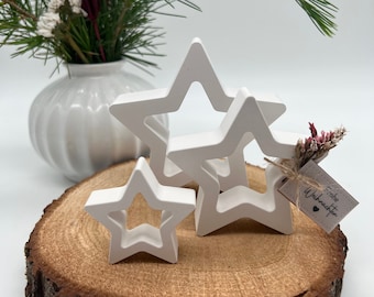 May include: Three white wooden star ornaments of varying sizes, arranged on a wooden slice. The largest star has a smaller star inside it. A small tag with the text "Frohe Weihnachten" is attached to the smallest star.