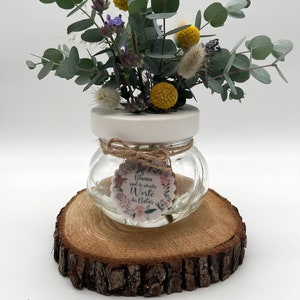Könnte beinhalten: Ein kleines Glasgefäß mit weißem Deckel und einem dekorativen Etikett mit der Aufschrift "Blumen sind die schönsten Worte der Natur" ist mit einem Strauß aus Eukalyptus, Lavendel und gelben Pomponblumen gefüllt. Das Gefäß steht auf einer Holzscheibe.