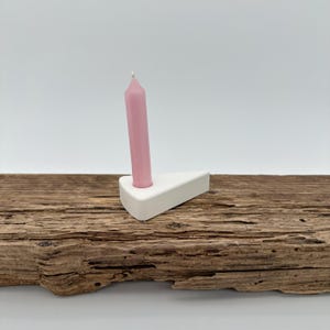 Könnte beinhalten: Ein weißer Kerzenhalter aus Keramik mit dreieckigem Design, der eine rosa Spitzkerze hält. Der Kerzenhalter steht auf einer verwitterten Holzoberfläche und schafft eine minimalistische Ästhetik. Die Kerze ist etwa 10 cm hoch.
