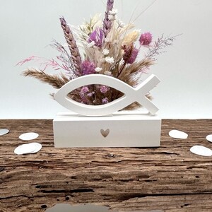 Könnte beinhalten: Weiße Holzvase in Fischform mit einem Herz-Ausschnitt, gefüllt mit getrockneten Blumen in Rosa-, Lila- und Weißtönen. Die Vase steht auf einer rustikalen Holzoberfläche.