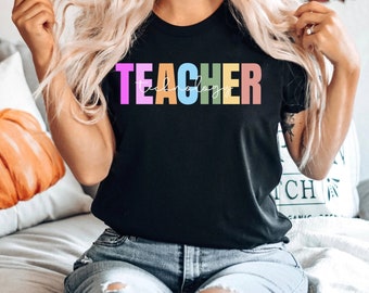 May include: A black t-shirt with the word "TEACHER" written in rainbow colors. The word "technology" is written in white script underneath the word "TEACHER".