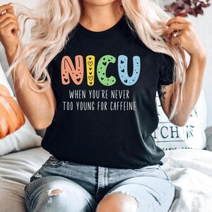 May include: Black t-shirt with a colorful "NICU" graphic and the text "When you're never too young for caffeine".
