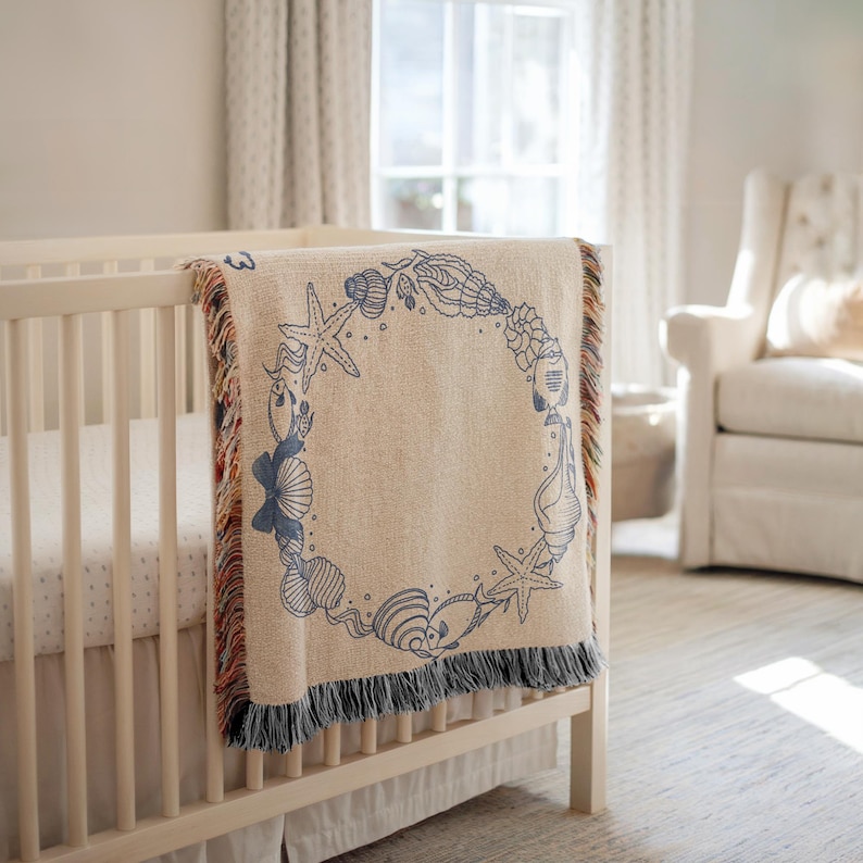 May include: A beige baby blanket with a blue seashell design draped over a white crib. The blanket features a circular pattern of starfish, shells, and seahorses. The edges have a fringe with blue, red, and brown threads.