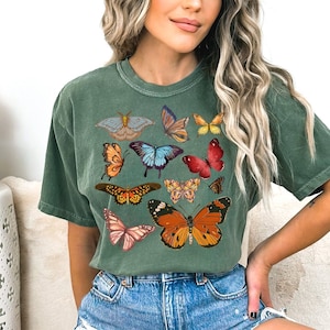 K&ouml;nnte beinhalten: Ein gr&uuml;nes T-Shirt mit verschiedenen bunten Schmetterlingsmotiven, die auf der Vorderseite aufgedruckt sind. Die Schmetterlinge sind in verschiedenen Gr&ouml;&szlig;en und Positionen angeordnet und erzeugen ein verspieltes und lebendiges Muster.