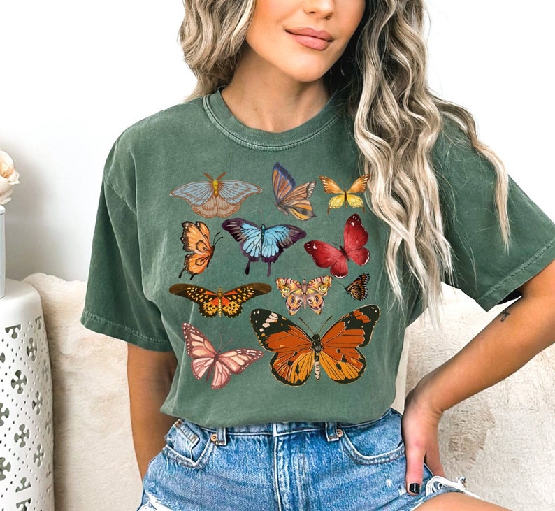 K&ouml;nnte beinhalten: Ein gr&uuml;nes T-Shirt mit verschiedenen bunten Schmetterlingsmotiven, die auf der Vorderseite aufgedruckt sind. Die Schmetterlinge sind in verschiedenen Gr&ouml;&szlig;en und Positionen angeordnet und erzeugen ein verspieltes und lebendiges Muster.