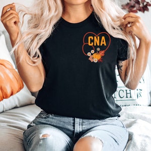 May include: Black t-shirt with a heart-shaped design containing the letters "CNA" in orange. The heart is decorated with a pumpkin, flowers, and fall leaves. The shirt is a crew neck.