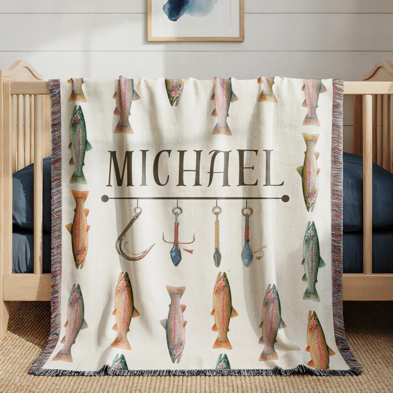 May include: Cream-colored woven throw blanket featuring a repeating pattern of watercolor fish in shades of orange, green, and brown. The name "MICHAEL" is displayed in a dark brown font above fishing-themed illustrations.