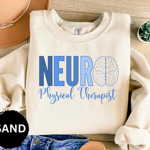 Puede incluir: Sudadera color arena con la palabra "NEURO" en letras azules, un gr&aacute;fico de cerebro y las palabras "Physical Therapist" en cursiva. La sudadera es de cuello redondo.