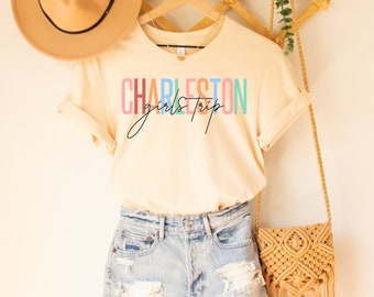 Könnte beinhalten: Ein beiges T-Shirt mit dem Text "CHARLESTON girls trip" in Regenbogenfarben. Das Shirt hängt an einem Holzbügel mit einem Strohhut und einer geflochtenen Tasche.