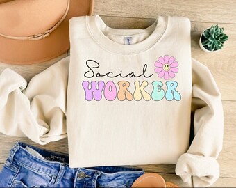 May include: A light beige sweatshirt with the text "Social Worker" in colorful, retro-style letters. A pink flower with a smiley face is above the text.
