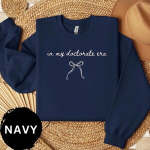 May include: Navy blue sweatshirt with the text "in my doctorate era" and a bow graphic in white. The word "NAVY" is in a black circle at the bottom. The sweatshirt is displayed on a woven surface.