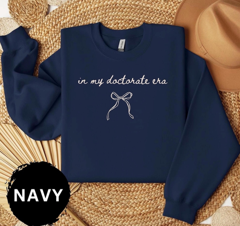 May include: Navy blue sweatshirt with the text "in my doctorate era" and a bow graphic in white. The word "NAVY" is in a black circle at the bottom. The sweatshirt is displayed on a woven surface.