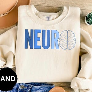 May include: A sand-colored sweatshirt with the word "NEURO" in blue block letters and a light blue brain graphic. The sweatshirt is folded on a wooden surface, with a small succulent plant in the background.