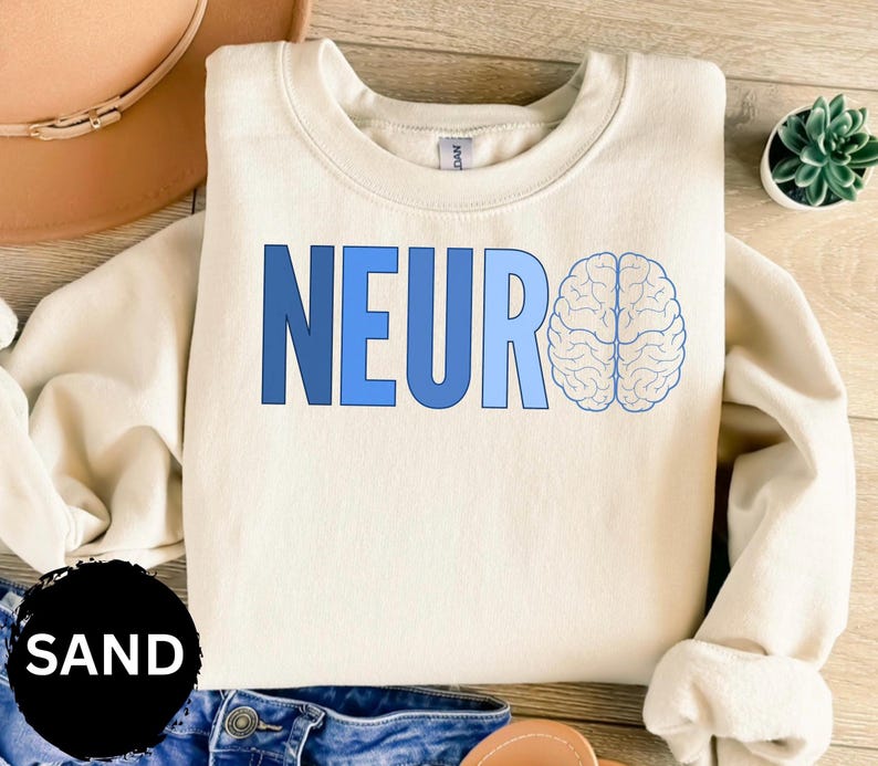 May include: A sand-colored sweatshirt with the word "NEURO" in blue block letters and a light blue brain graphic. The sweatshirt is folded on a wooden surface, with a small succulent plant in the background.