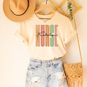 May include: A beige t-shirt with the word "NURSE" printed in colorful vertical stripes. The name "Katelyn" is written in cursive below the word "NURSE". The shirt is hanging on a wooden hanger with a straw hat and a woven bag.