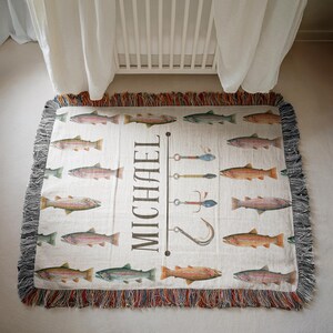 May include: Cream-colored woven throw blanket featuring a pattern of colorful fish and fishing lures. The name "MICHAEL" is printed vertically in black. The blanket has a fringed edge in shades of red, brown, and gray.