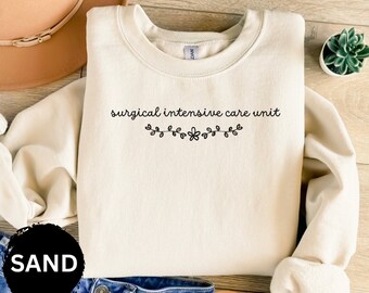 May include: A cream-colored sweatshirt with the text "surgical intensive care unit" in black lettering with a small floral design underneath. The sweatshirt is laid out on a wooden surface.