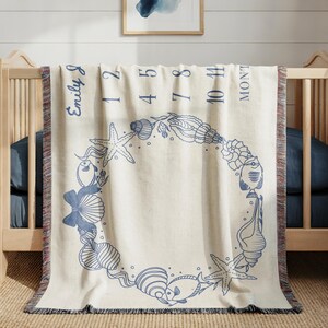 May include: Cream-colored baby milestone blanket with a blue seashell and starfish wreath design. The blanket has the name "Emily J" and numbers 1, 2, 4, 5, 7, 8, 10, and 11, with the word "MONTHS" printed on it. The blanket is fringed.