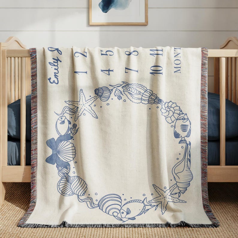May include: Cream-colored baby milestone blanket with a blue seashell and starfish wreath design. The blanket has the name "Emily J" and numbers 1, 2, 4, 5, 7, 8, 10, and 11, with the word "MONTHS" printed on it. The blanket is fringed.