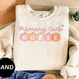 May include: A cream-colored sweatshirt with the text "Primary Care Nurse" in pink and orange lettering. The text is surrounded by five pink and orange pumpkins with the letters "NURSE" spelled out on them. The sweatshirt is on a wooden surface.