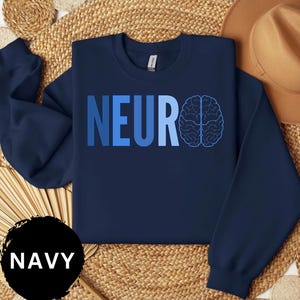 May include: Navy blue sweatshirt with the word "NEURO" in gradient blue letters and a brain graphic. The word "NAVY" is in a black circle at the bottom left. The sweatshirt is on a woven surface.